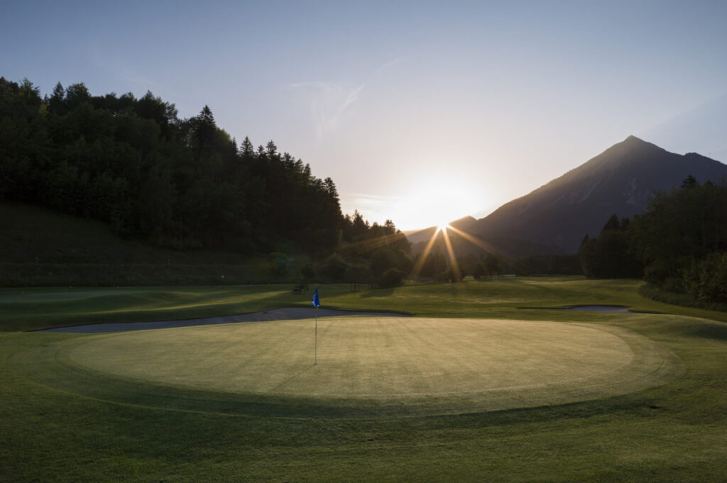 Golfplatz in den Bergen bei Sonnenaufgang mit beleuchtetem Green und Fahne vor alpiner Kulisse.
