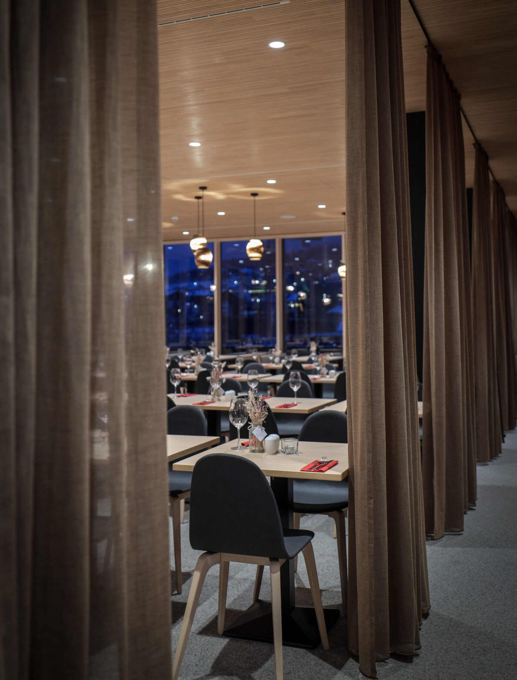 Blick durch Raumteiler in den abendlich beleuchteten Restaurantbereich des Food Market by AVES Savognin mit gedeckten Tischen und stimmungsvollem Licht.