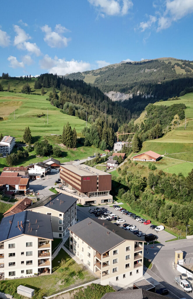 Drohnenaufnahme Hotel AVES Savognin von oben – Hotelgebäude mit Parkplatz, Bergbahnen und alpiner Landschaft im Hintergrund