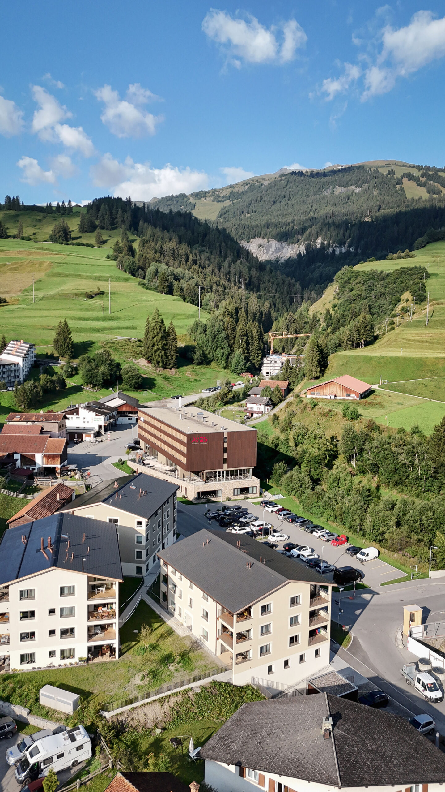 Luftaufnahme von Savognin mit dem AVES Hotel im Zentrum, umgeben von grünen Wiesen und sanften Bündner Bergen im Sommer
