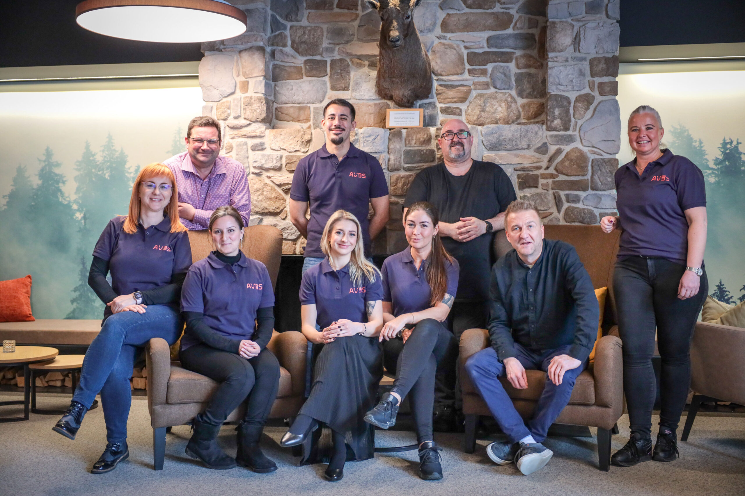 Das Team des AVES Hotel Savognin posiert gemeinsam vor einem Steinkamin in der Hotel-Lounge.