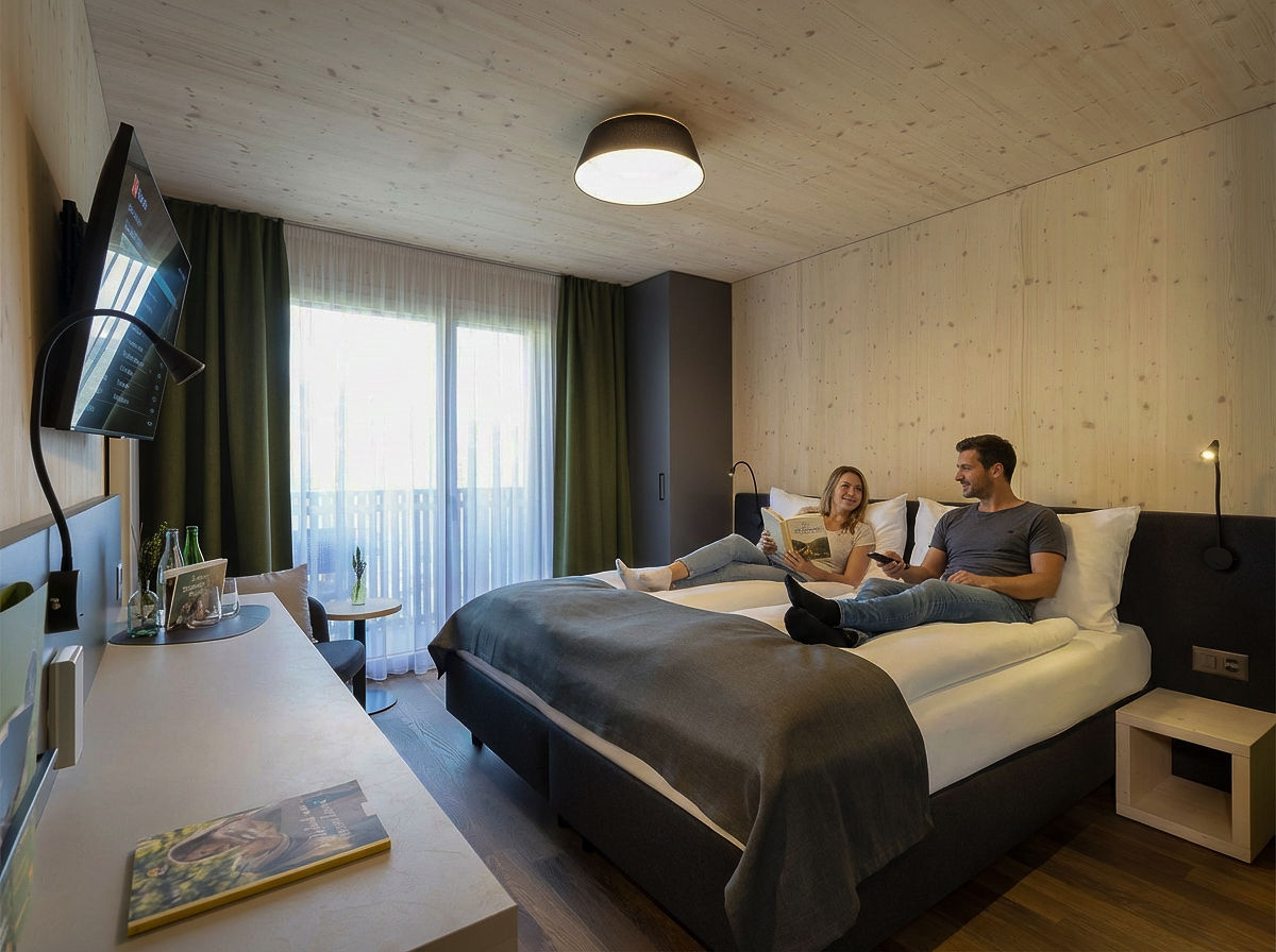 Pärchen entspannt auf dem Bett im modernen Doppelzimmer des AVES Savognin mit Holzverkleidung, Balkon und grossem TV