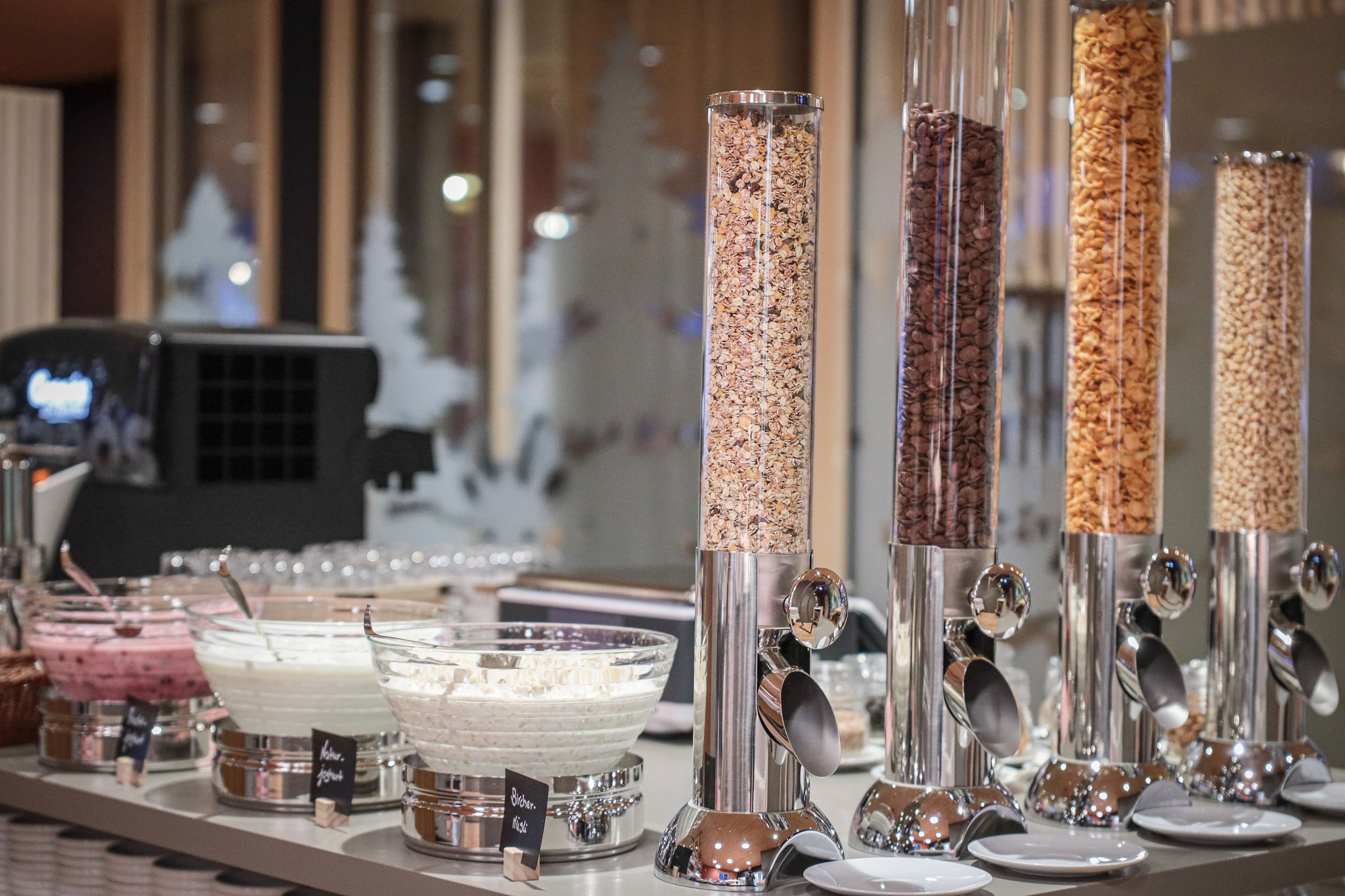Getreidebar mit verschiedenen Müesli-Sorten in Glasspendern sowie Naturjoghurt und Bircher-Müesli in Schalen