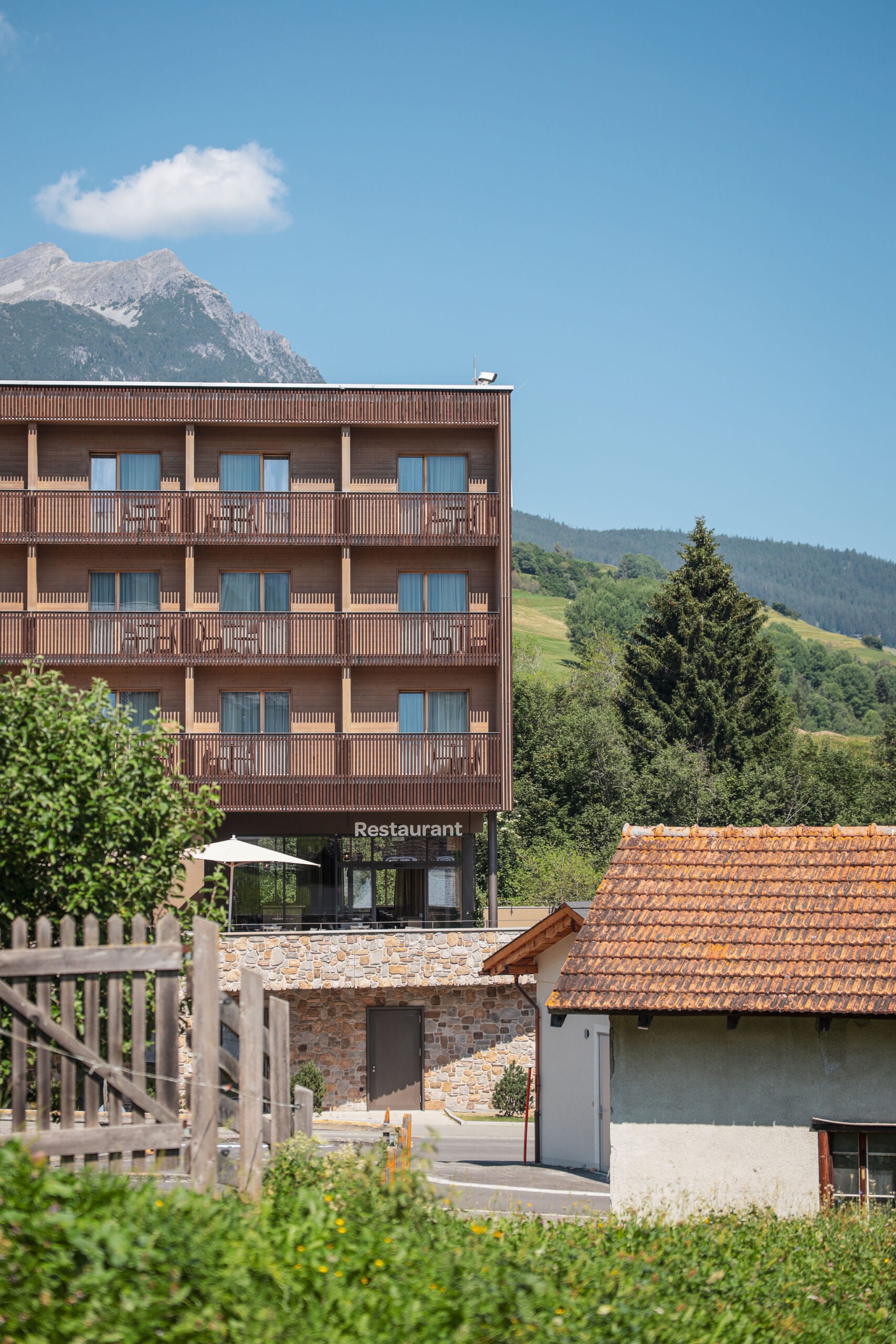 Aussenansicht des AVES Hotels Savognin mit Balkonen und Restaurant-Schriftzug, umgeben von grünen Hügeln, Bergpanorama und einem traditionellen Bündner Steinhaus im Vordergrund.
