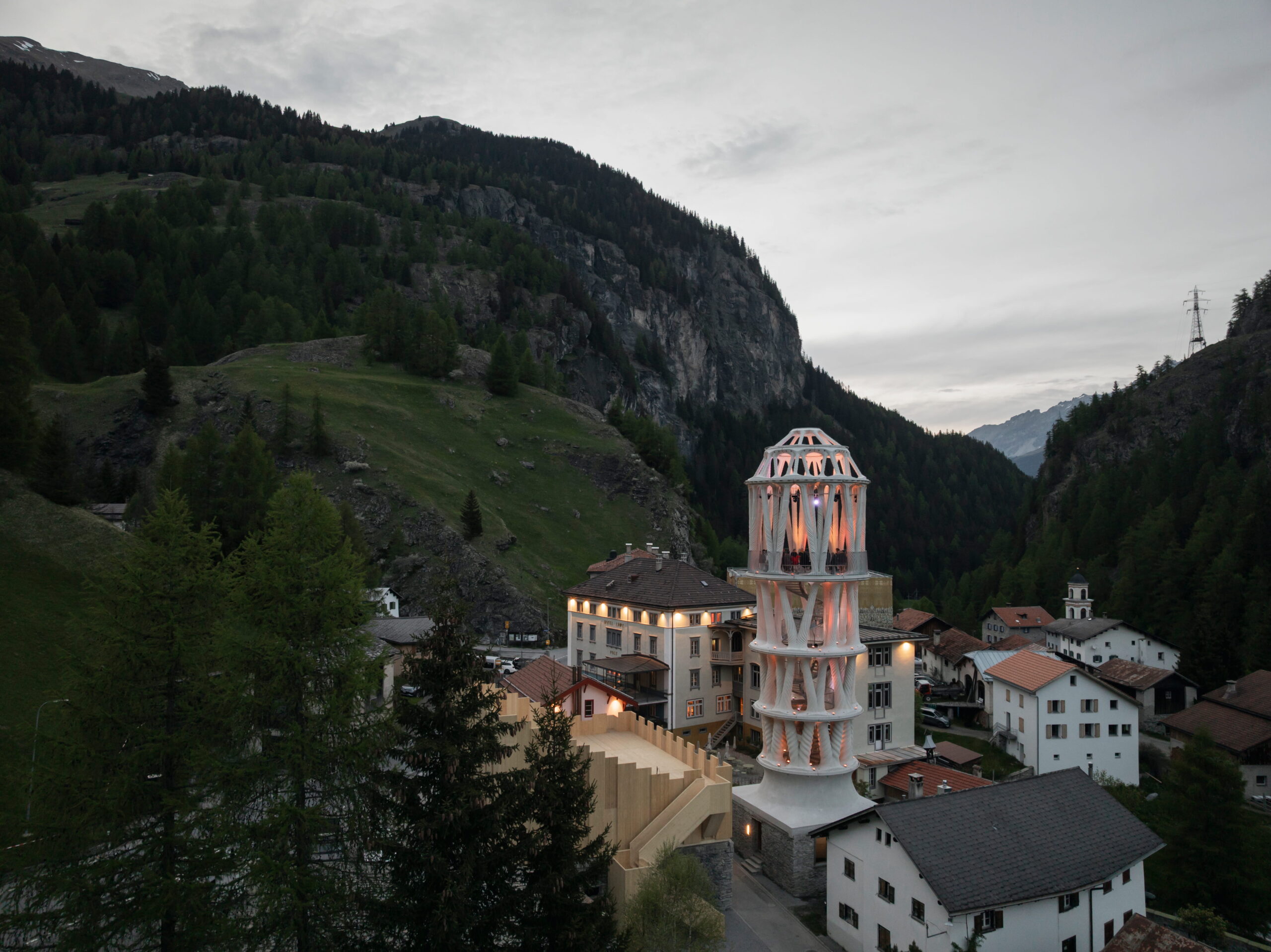 Der illuminierte Weisse Turm in Mulegns bei Abenddämmerung, eingebettet ins Dorfbild des Vals Surses mit bewaldeten Berghängen im Hintergrund – Ausflugsziel der geführten E-Bike Tour ab AVES Hotel Savognin