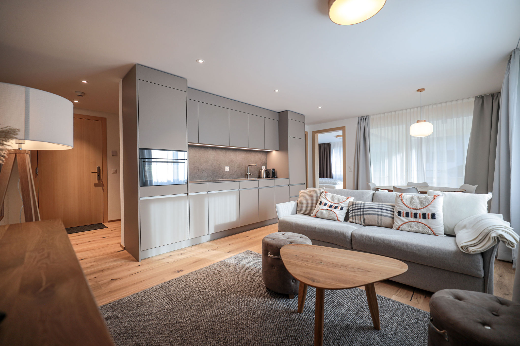 Offene Wohnküche mit grauen Küchenschränken, Kühlschrank und Kaffeemaschine links, davor ein graues Sofa mit gemusterten Kissen, runder Holztisch und zwei Sitzhocker auf grauem Teppich, Holzboden, im Hintergrund durch eine Tür Schlafzimmer und Essbereich mit Vorhängen sichtbar.