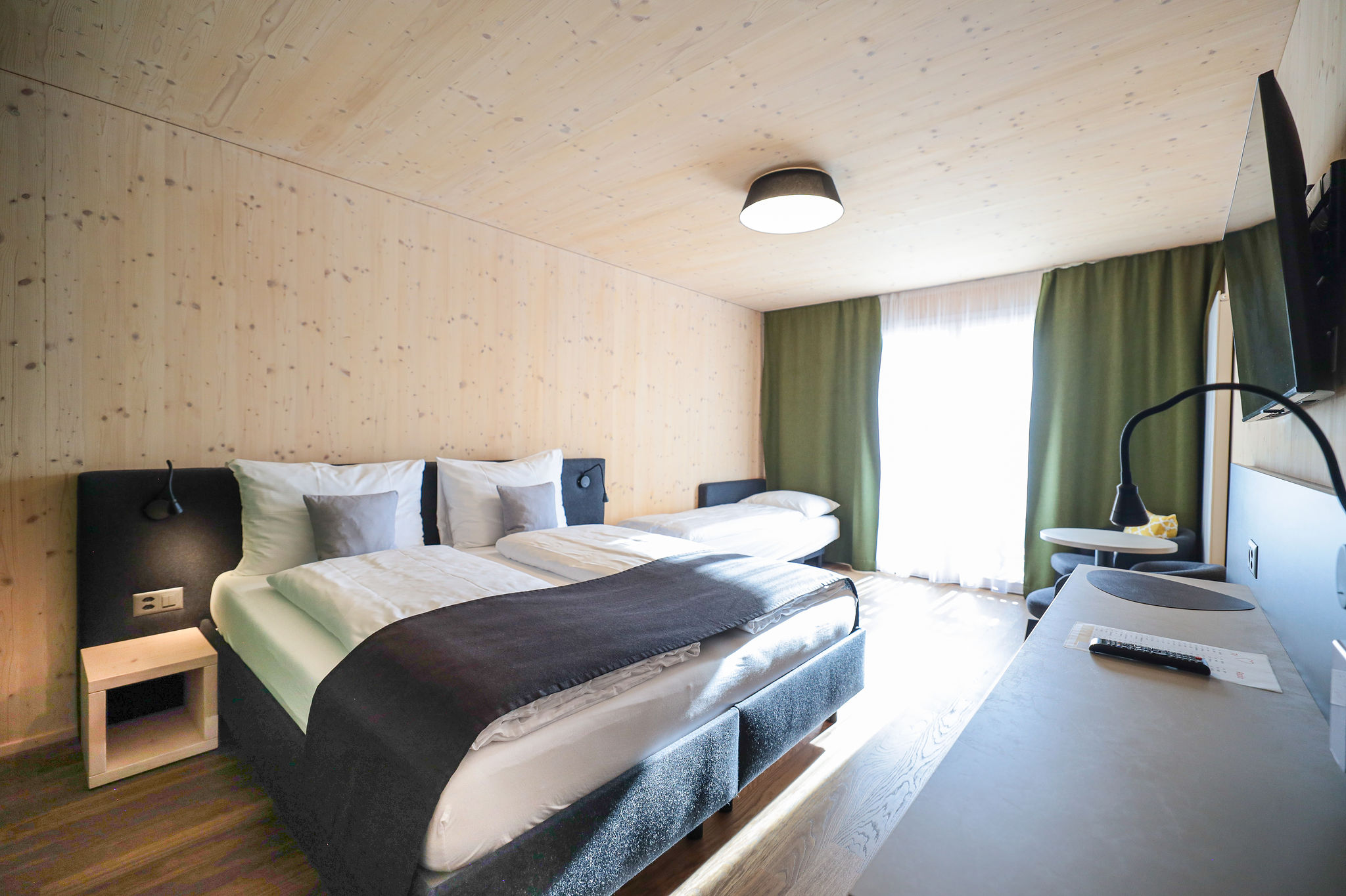 Hotelzimmer im Aves Savognin mit Holzverkleidung an Wänden und Decke, Doppelbett mit dunkelgrauem Polsterkopfteil und zusätzlichem Einzelbett, dunkelgrüne Vorhänge vor der Balkontür, rechts Schreibtisch mit TV und Leselampe, im Hintergrund kleiner Sitzbereich mit Rundtisch.