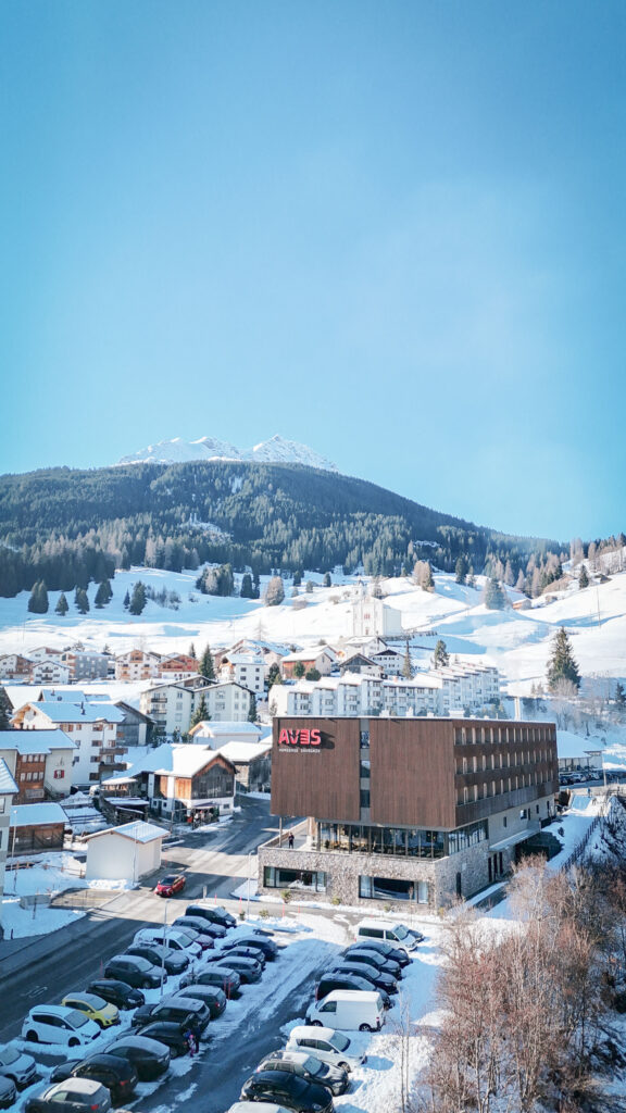 Das AVES Homebase Savognin mit Holzfassade und Steinsockel im verschneiten Dorf, dahinter die weisse Kirche am Hang, Nadelwald und ein verschneiter Gipfel.