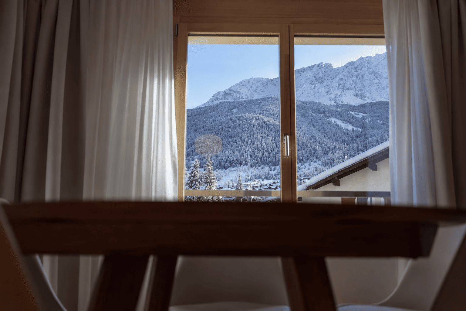 Blick über einen Holztisch im Apartment durch ein Fenster mit Holzrahmen und hellen Vorhängen auf verschneiten Nadelwald und felsige, schneebedeckte Berggipfel.
